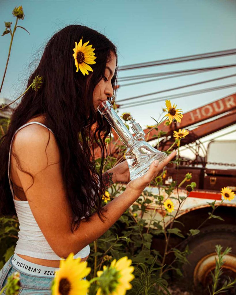 Young woman surrounded by flowers taking a hit from a glass bong
